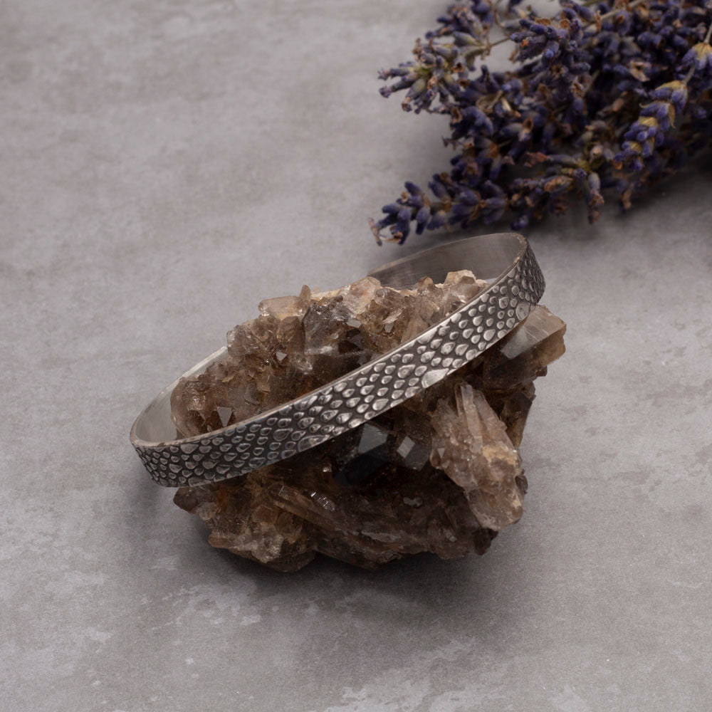 Sterling silver cuff bracelet with a raindrop texture, displayed on an amethyst crystal with lilac sprigs on the background.
