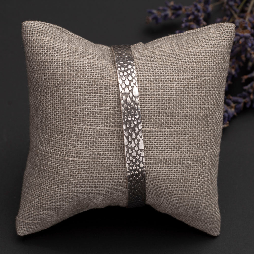 Sterling silver cuff bracelet with a raindrop texture, displayed on soft fabric pillow with dried lilac for decoration.