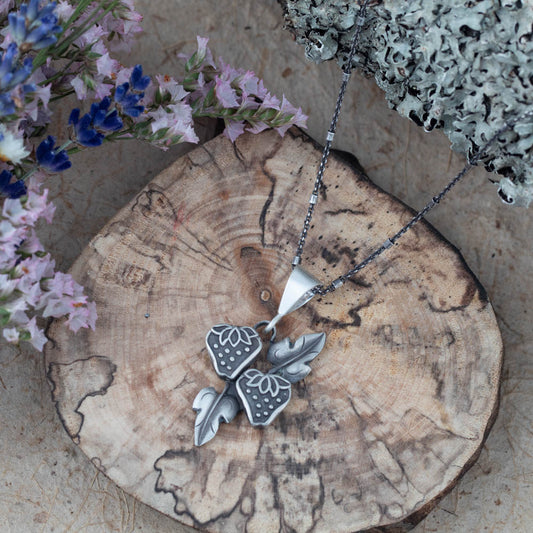 Sterling silver strawberry and leaf pendant by Divella Designs, handmade in Vermont with patina finish on an 18" chain, photographed on wood with lichen and dried flowers