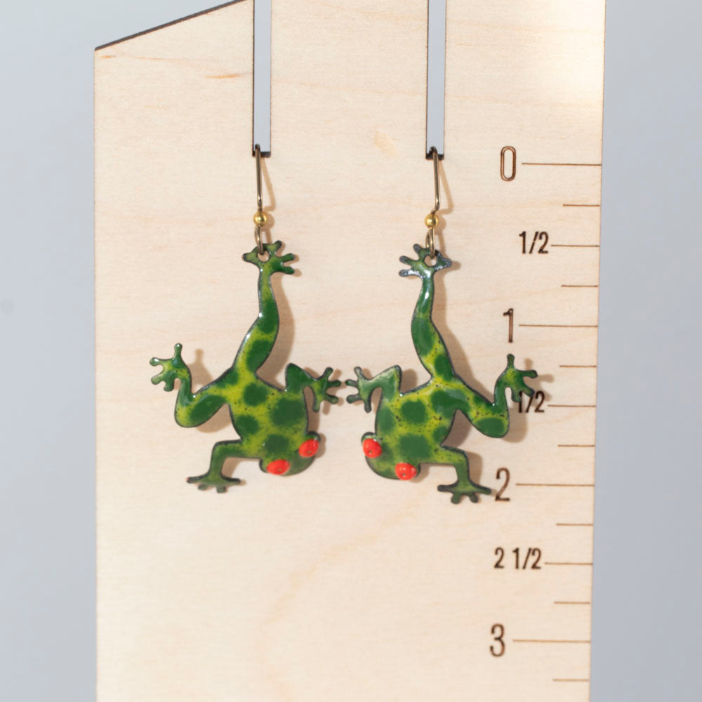 Playful green frog-shaped enamel earrings with bright red eyes, handcrafted and displayed on wooden ruler for scale– Enchanted Frog design by Divella Design