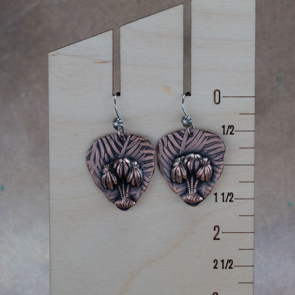 Handcrafted copper earrings with sculpted palm trees layered over embossed leaf-patterned backdrops, tropical artisan jewelry by Divella Designs – displayed on a wooden ruler for scale.