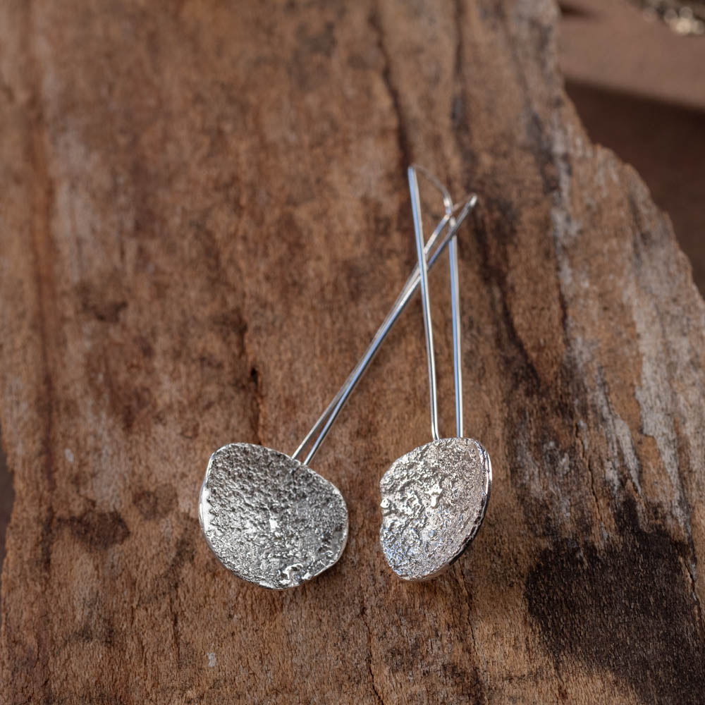 Sterling silver dangle earrings with textured, reticulated surfaces hanging from long ear wires, displayed against raw bark — handmade by Divella Designs Jewelry.