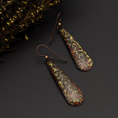 Long teardrop-shaped enamel earrings with a speckled pattern in shades of yellow, red, and black, displayed on a dark grey background with sprigs of autumn greenery.