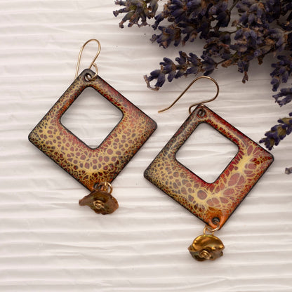 Diamond-shaped enamel earrings with a yellow and red organic crackle pattern, accented by golden woodland-inspired keishi pearl dangles, displayed on white textured paper with sprigs of lilac.
