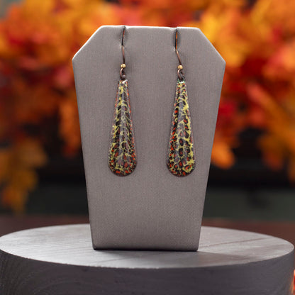 Long teardrop-shaped enamel earrings with a speckled pattern in shades of yellow, red, and black, displayed upright on a gray stand with a blurred autumn foliage background.
