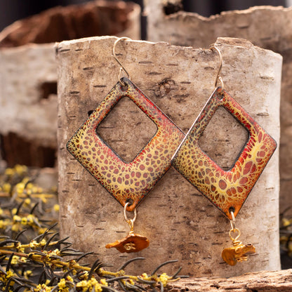 Diamond-shaped enamel earrings with a yellow and red organic crackle pattern, accented by golden keishi pearl dangles, displayed on birch bark with yellow wildflowers.