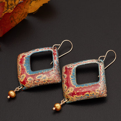 Diamond-shaped enamel earrings in rich red, blue, and golden tones with a cut-out center and warm golden bead dangles, displayed on a dark gray surface with autumn leaves in the background.