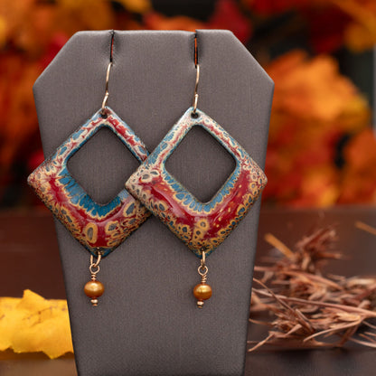 Diamond-shaped enamel earrings in rich red, blue, and golden tones with a cut-out center and warm golden bead dangles, displayed on a gray stand with autumn leaves in the background.
