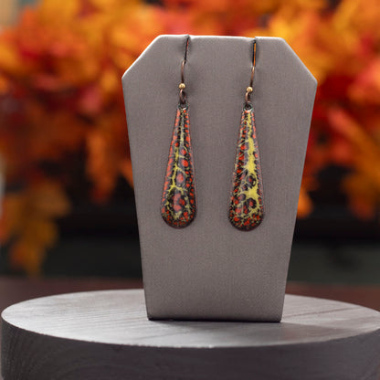 Long teardrop-shaped enamel earrings in fiery shades of red, yellow, and black with organic patterns, displayed on a gray stand with a blurred autumn leaf background.