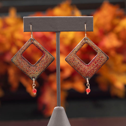 Diamond-shaped enamel earrings in rich orange and red tones with textured speckled pattern, featuring small red gemstone dangles, displayed against a fall foliage background.