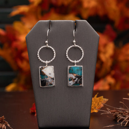 Sterling silver dangle earrings with twisted circle accents and rectangular composite stones made from white buffalo stone, Kingman turquoise, and bronze, displayed on a gray stand with colorful autumn leaves in the background.
