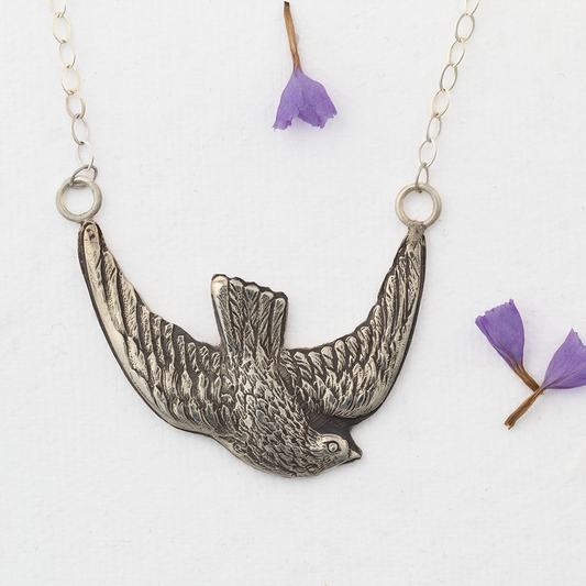 Sterling silver bird in flight pendant necklace with detailed feather texture, displayed on white background with scattered purple flower petals.