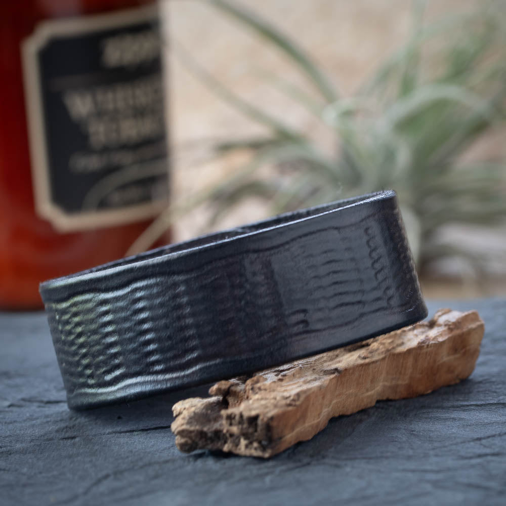 Black textured leather cuff on a wooden stand with a blurred bottle and plant in the background