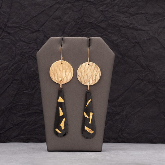 Elegant black enamel teardrop earrings featuring 24k gold flake accents and hammered brass discs, displayed on a gray stand with textured dark background.