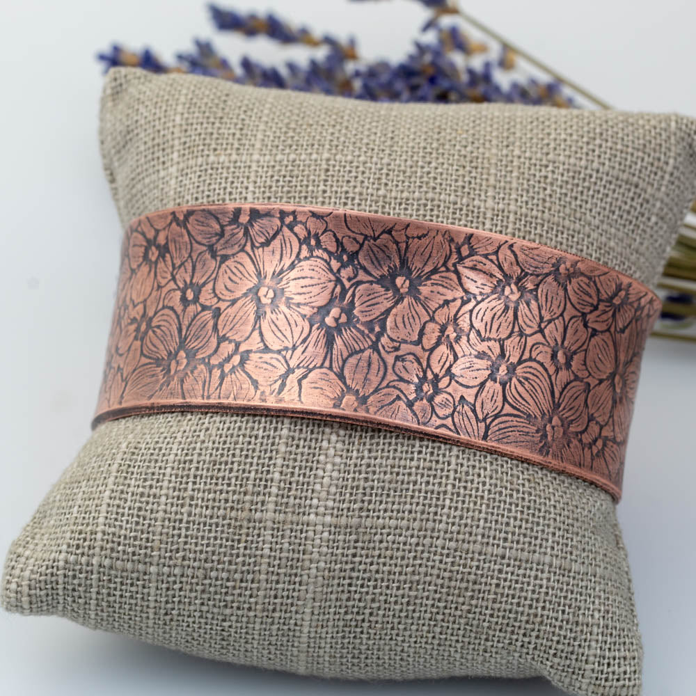 Anticlastic copper cuff bracelet with detailed floral pattern displayed on linen pillow.