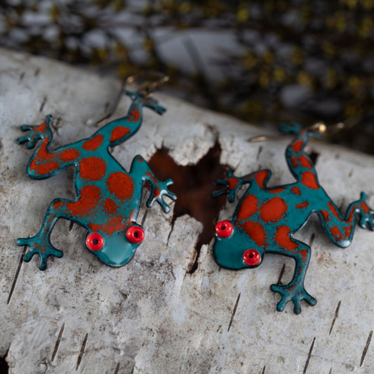 Handcrafted enameled copper frog earrings in bold red and teal with bright red eyes, tree frog design displayed on rustic birch bark – artisan nature jewelry by Divella Design