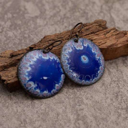 Round dangle earrings with a bold blue enamel center, surrounded by a delicate purple and white petal-like border pattern, displayed on a rustic piece of wood.