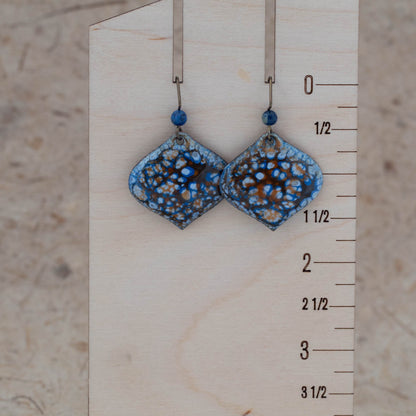 Tidal Pool Enamel Earrings – handcrafted diamond-shaped copper earrings with ocean blue, white, and rust-toned enamel glaze, accented by blue gemstone beads. Displayed on rustic birch bark with wildflowers.