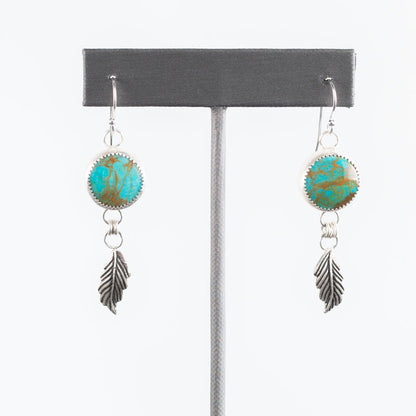 Sterling silver earrings with round bezel-set turquoise stones and dangling feather charms, handcrafted Southwestern-inspired design by Divella Designs – shown on white background.