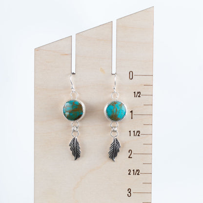 Sterling silver earrings with round bezel-set turquoise stones and dangling feather charms, handcrafted Southwestern-inspired design by Divella Designs – shown on wooden ruler for scale.