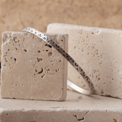 Handmade sterling silver botanical bangle with small floral impressions, photographed on stacked stone blocks with neutral tones and lavender in the background.