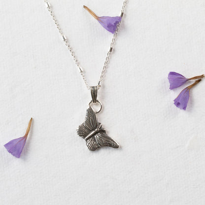 Delicate sterling silver butterfly pendant necklace with fine wing detailing, displayed on a white background with scattered purple flower petals.