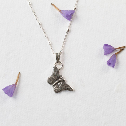 Delicate sterling silver butterfly pendant necklace with fine wing detailing, displayed on a white background with scattered purple flower petals.