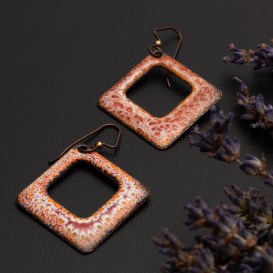 Square copper enamel earrings in warm earthy tones of rust, orange, and cream with a speckled crackle pattern, displayed on a dark surface with dried lavender sprigs.