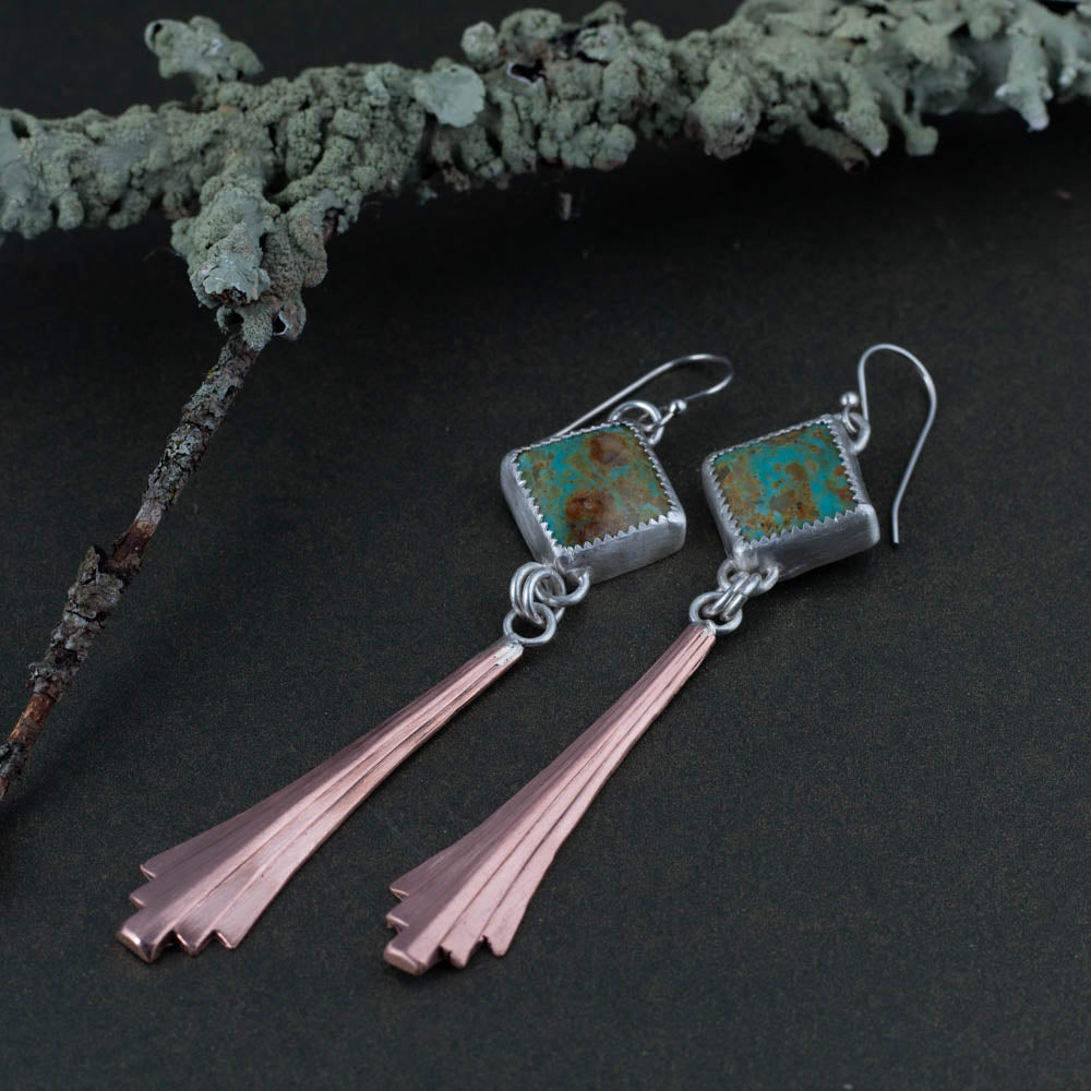 Pair of earrings with square kingman turquoise stones and copper-like extensions on a dark surface.