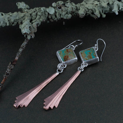 Pair of earrings with square kingman turquoise stones and copper-like extensions on a dark surface.