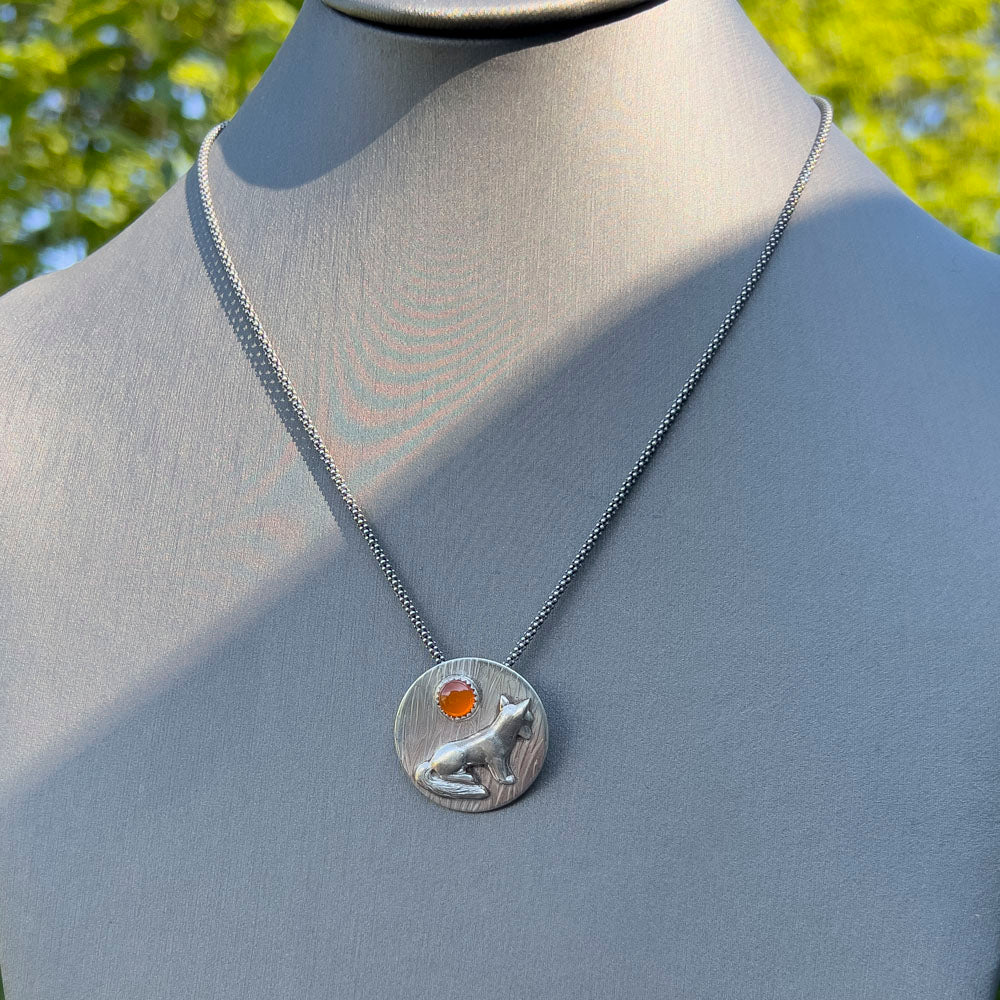 A silver pendant necklace featuring a fox and a carnelian stone, displayed on a gray mannequin bust with a blurred green outdoor background.