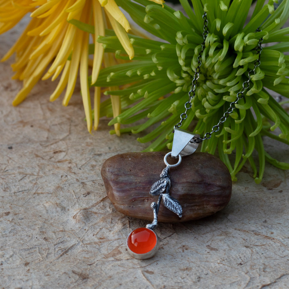 Handmade sterling silver mandarin orange pendant with a bright AAA-grade carnelian stone and oxidized leaves displayed with flowers and a smooth stone