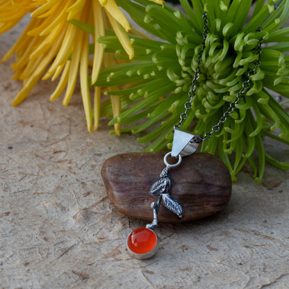 Handmade sterling silver mandarin orange pendant with a bright AAA-grade carnelian stone and oxidized leaves displayed with flowers and a smooth stone