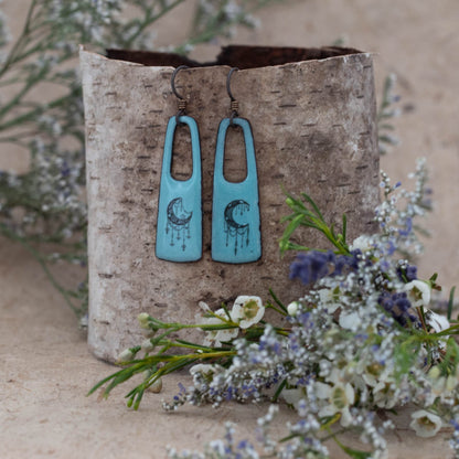 Handmade turquoise blue enamel earrings with celestial crescent moon and star illustrations in black. Bohemian-style dangle earrings with open rectangular design, displayed on birch bark with wildflowers – artisan jewelry by Divella Designs.