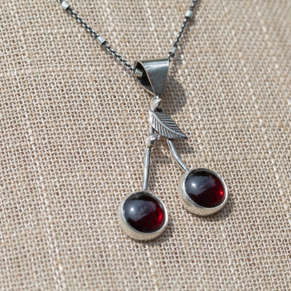 Close view of a handmade garnet cherry necklace with sterling silver stems and a detailed silver leaf. Photo taken outside,