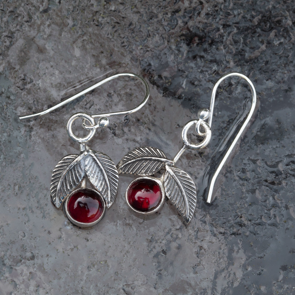 Garnet and sterling silver cherry earrings with textured silver leaves and French hook ear wires on a gray stone background.