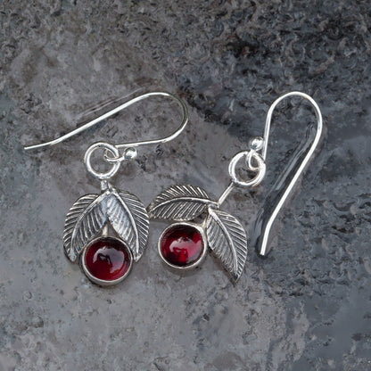 Garnet and sterling silver cherry earrings with textured silver leaves and French hook ear wires on a gray stone background.