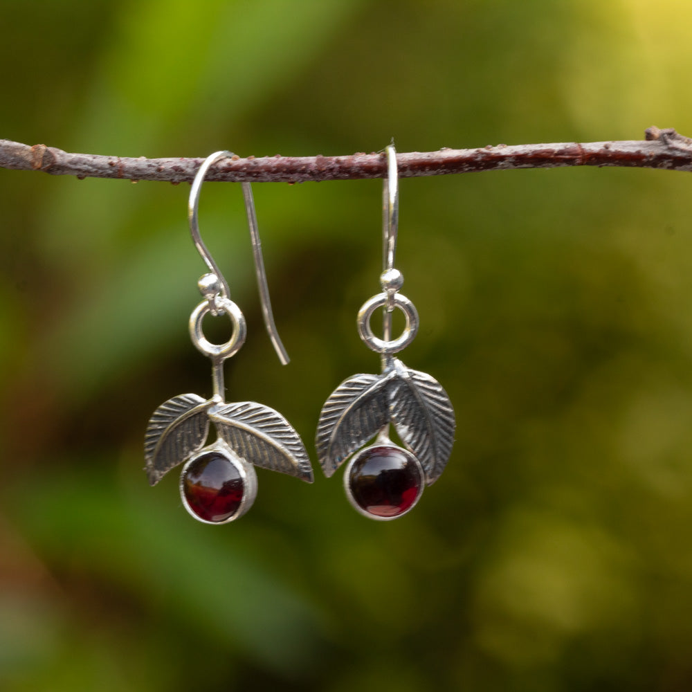 Sterling silver cherry earrings with dark red garnet stones and textured leaves hanging from a branch against a green background.