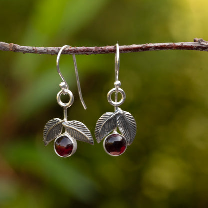 Sterling silver cherry earrings with dark red garnet stones and textured leaves hanging from a branch against a green background.