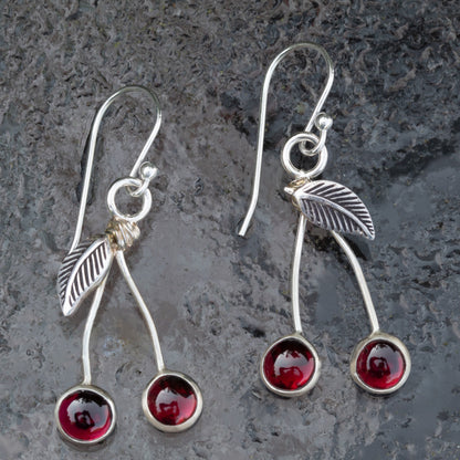 Garnet and sterling silver cherry earrings with curved stems, textured leaves, and French hook ear wires on a gray stone background.