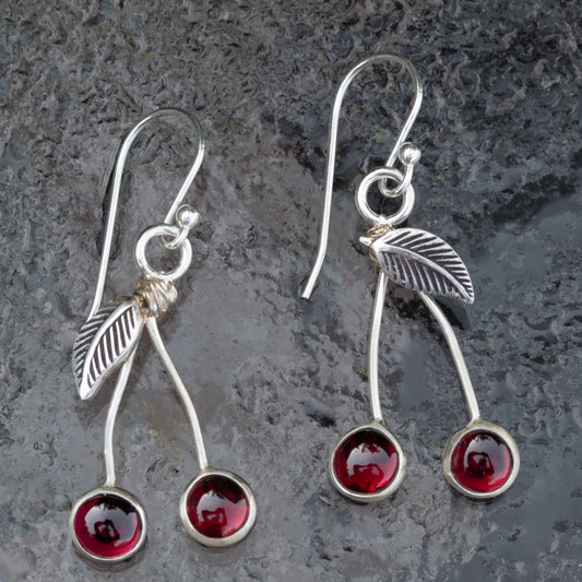Garnet and sterling silver cherry earrings with curved stems, textured leaves, and French hook ear wires on a gray stone background.
