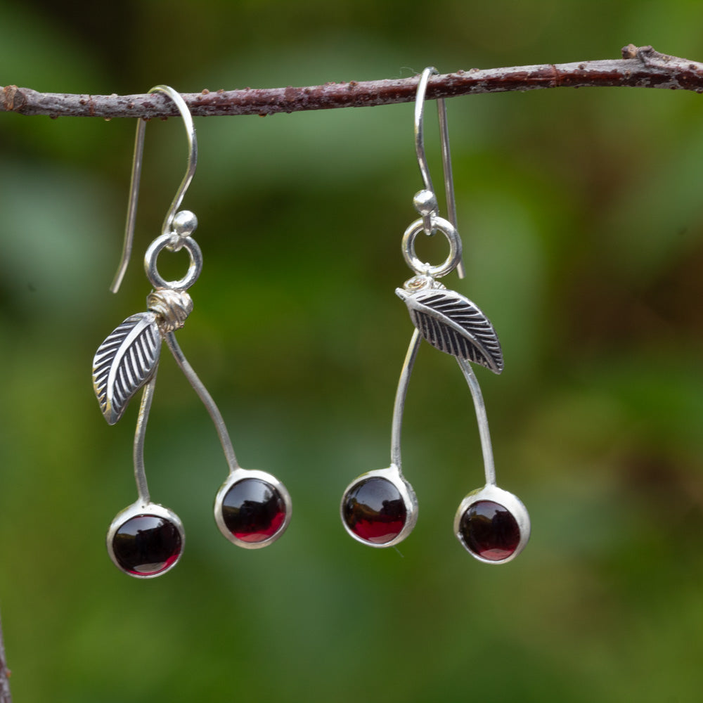 Sterling silver cherry earrings with dark red garnet stones and textured leaves hanging from a branch against a green background.