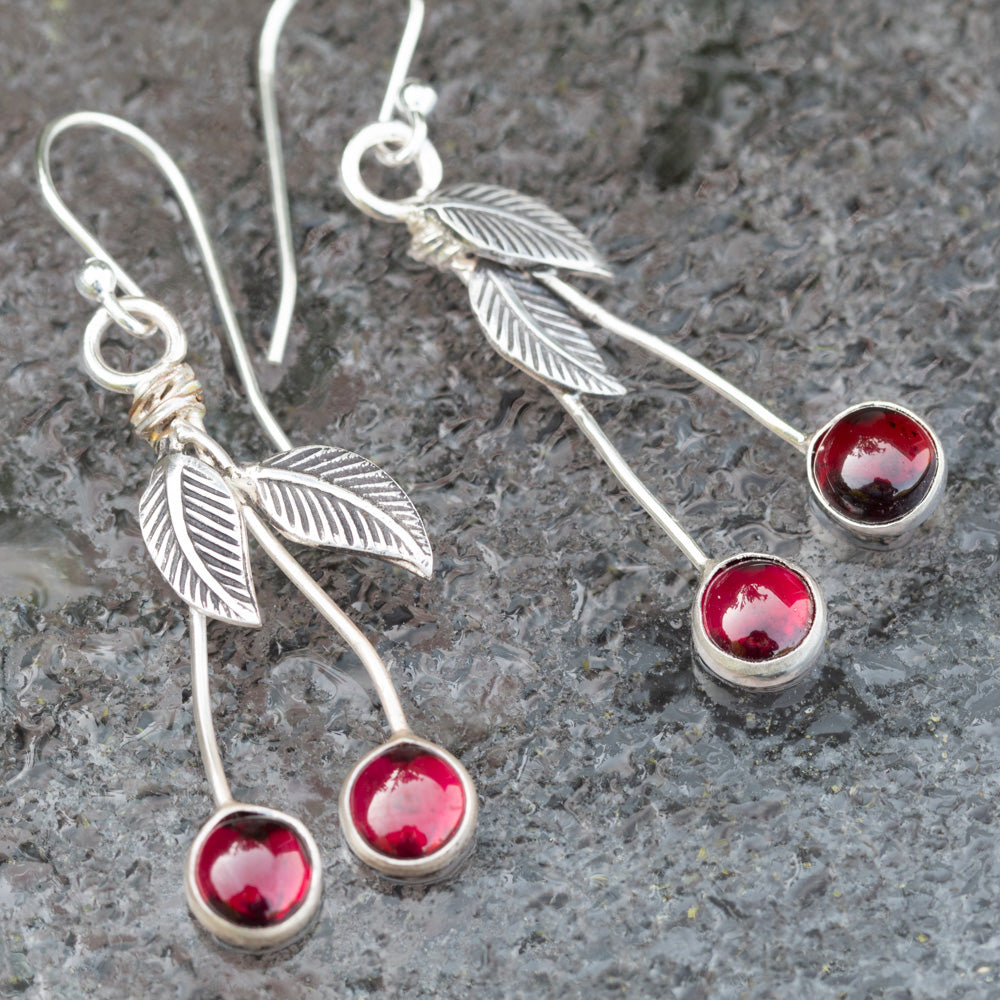 Garnet and sterling silver cherry drop earrings with textured leaves and long silver stems on a gray stone background.
