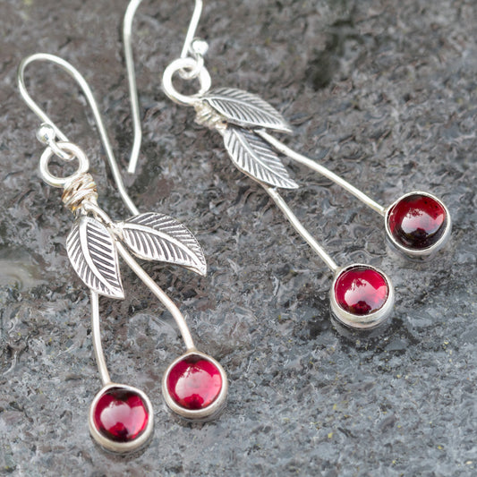 Garnet and sterling silver cherry drop earrings with textured leaves and long silver stems on a gray stone background.