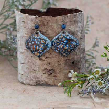 Coastal Drift Enamel Earrings – handmade diamond-shaped copper earrings with ocean-inspired blue, white, and rust enamel patterns, accented with natural stone beads. Displayed on birch bark with wildflowers.