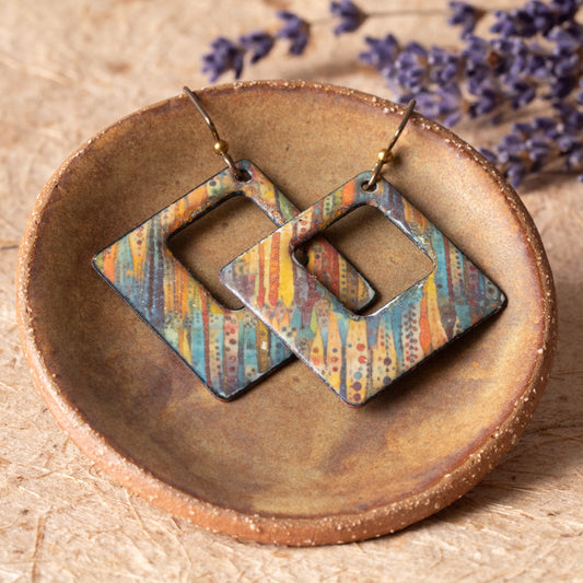 Colorful open square enamel earrings with striped blue, yellow, orange and rust patterns displayed in a handmade pottery dish.
