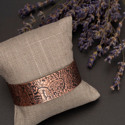 Copper magic mushroom designed cuff is pictured on a jewelry cushion lying on a gray background with sprigs of lavender.