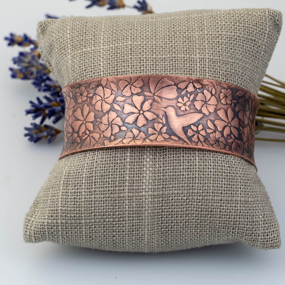 Copper bracelet with hummingbird floral patterns on a textured beige pillow