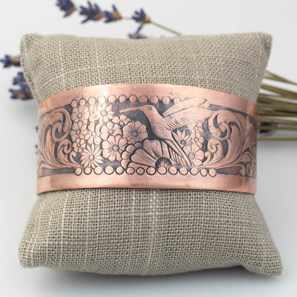 Copper bracelet with intricate hummingbird and floral designs on a textured fabric pillow