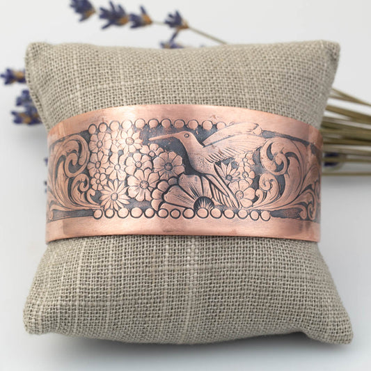 Copper bracelet with intricate hummingbird and floral designs on a textured fabric pillow